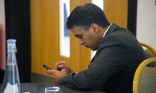 Man in a suit sitting at a table, intently using a smartphone, with a water bottle and glass in the foreground.