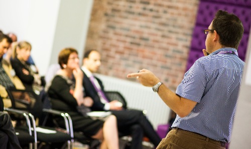 Man in a checkered shirt speaking to a seated audience in a modern conference room with a brick and purple-accented wall.
