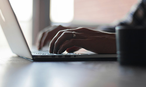 Close-up of hands typing on a laptop keyboard near a window, with a metal ring visible on one finger.